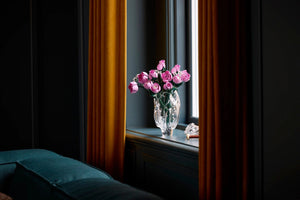 Bouquet of pink roses in a clear vase on a windowsill with dark curtains.