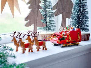 Toy Santa Claus in a sleigh with reindeer on a windowsill with decorative trees in the background.